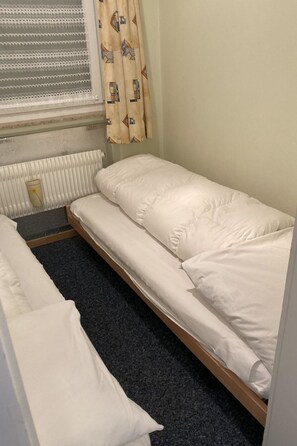 Ein kleines Zimmer mit einem Einzelbett, einem Heizkörper, einem Fenster mit Jalousien und einem gemusterten Vorhang.