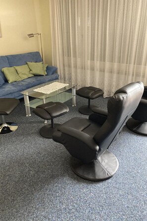 Ein Raum mit einem schwarzen Massagesessel, einem Glastisch, einem blauen Sofa mit grünen Kissen und einer Stehlampe.