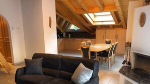 Uma sala de estar moderna com um sofá de couro preto, uma área de jantar com uma mesa redonda e cadeiras, e uma cozinha com armários de madeira e um claraboia.