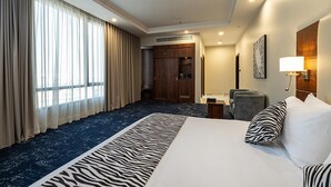 Quarto de hotel com cama grande, tapete estampado com padrão de zebra, escrivaninha, cadeira, guarda-roupa e uma janela ampla com cortinas.