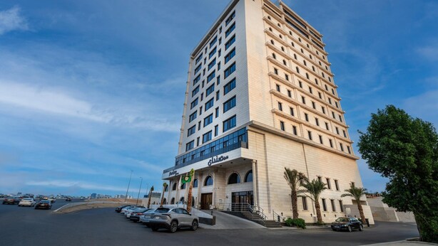 Hotel com vários andares e design moderno, com entrada destacada, escada e área de estacionamento à frente.