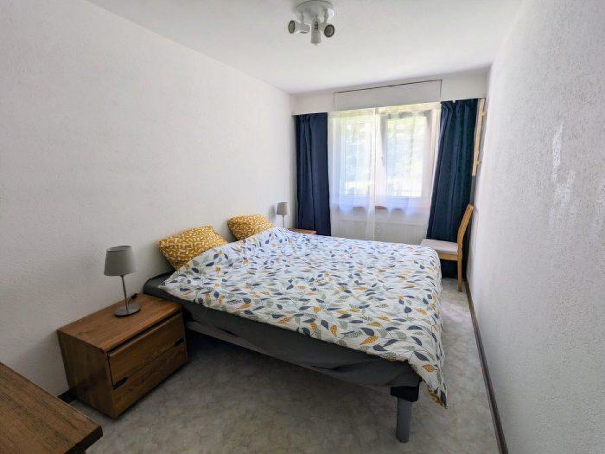 Ein Schlafzimmer mit Bett, Nachttisch, Lampe und einem Fenster mit Vorhängen.