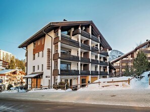 Et hotel med flere etager og balkoner, med en skibakke i baggrunden og en klar himmel.