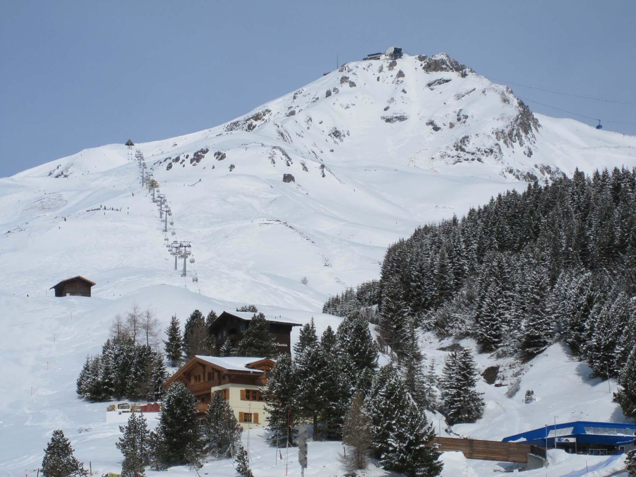 Un refugio de montaña con un telesilla, rodeado de árboles cubiertos de nieve y un cielo despejado.
