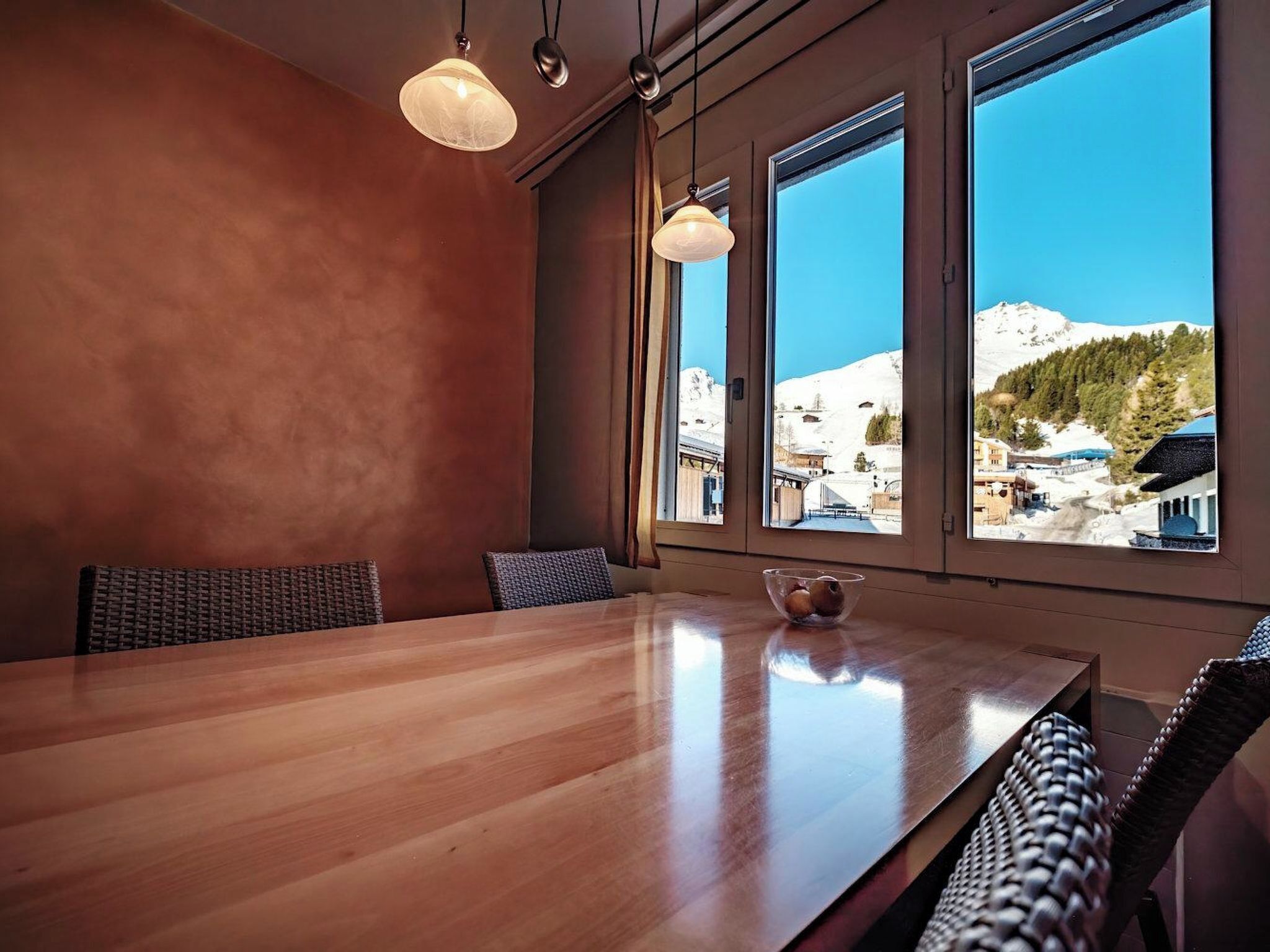 A wooden dining table with chairs, large windows with a view of a snowy mountain, and pendant lights.