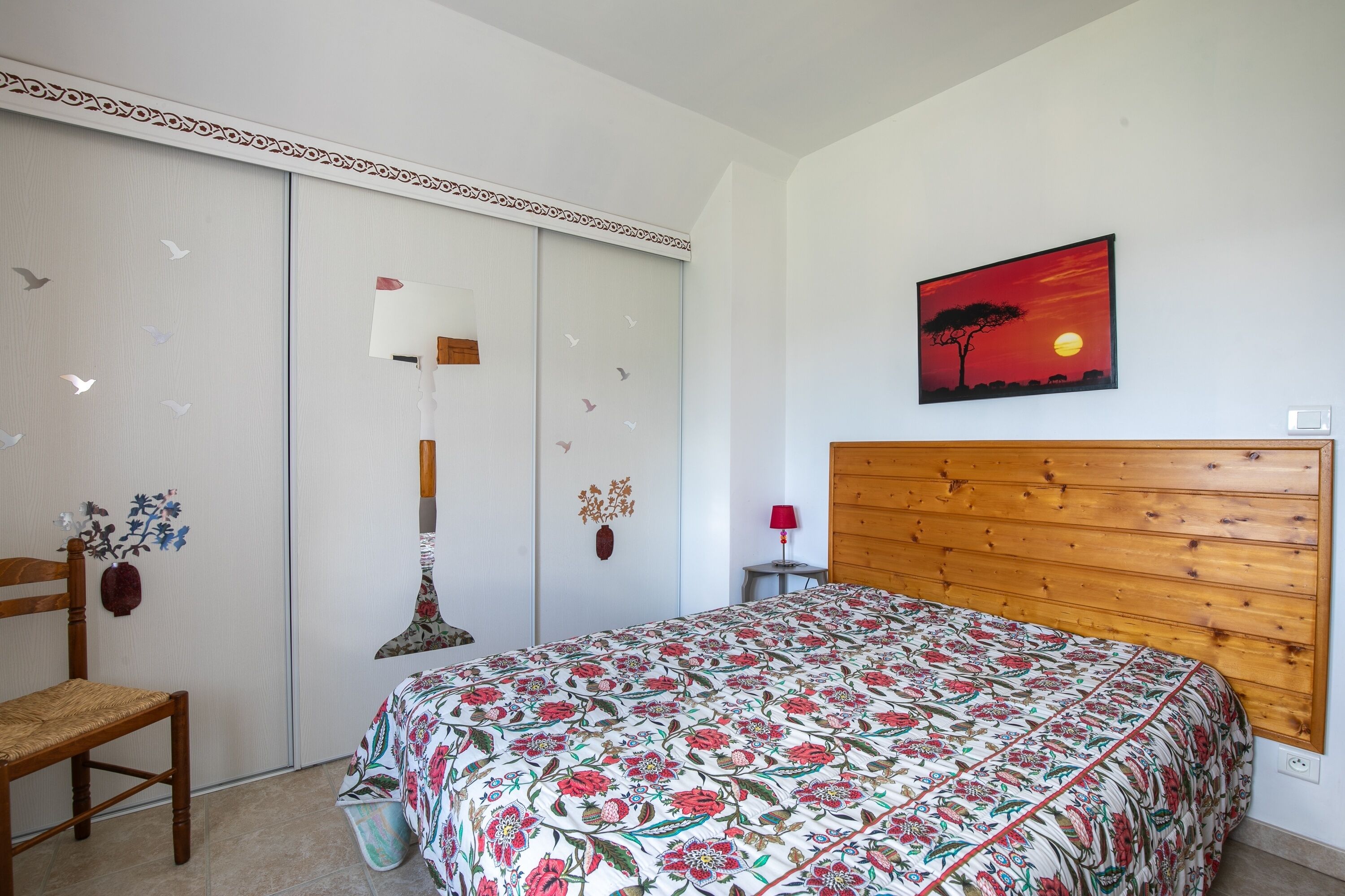 Un dormitorio con cabecera de madera, colcha estampada con flores, lámpara de pared y una pintura de atardecer.