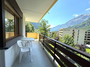 A balcony with a table and chair, overlooking a mountain landscape and a building labeled 'FORTUNA'. - Royal 32 # 6-bed apartment, Dusche, WC 6-Bettwohnung, Dusche, WC by Interhome (Leukerbad)