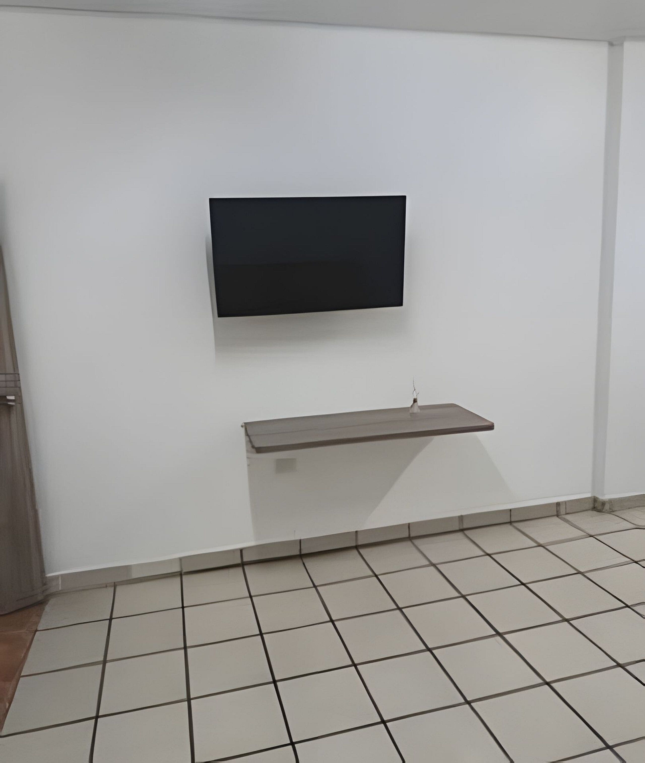A room with a flat-screen TV mounted on the wall, a small shelf below it, and a tiled floor.