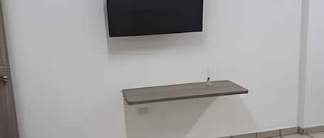 A room with a flat-screen TV mounted on the wall, a small shelf below it, and a tiled floor.