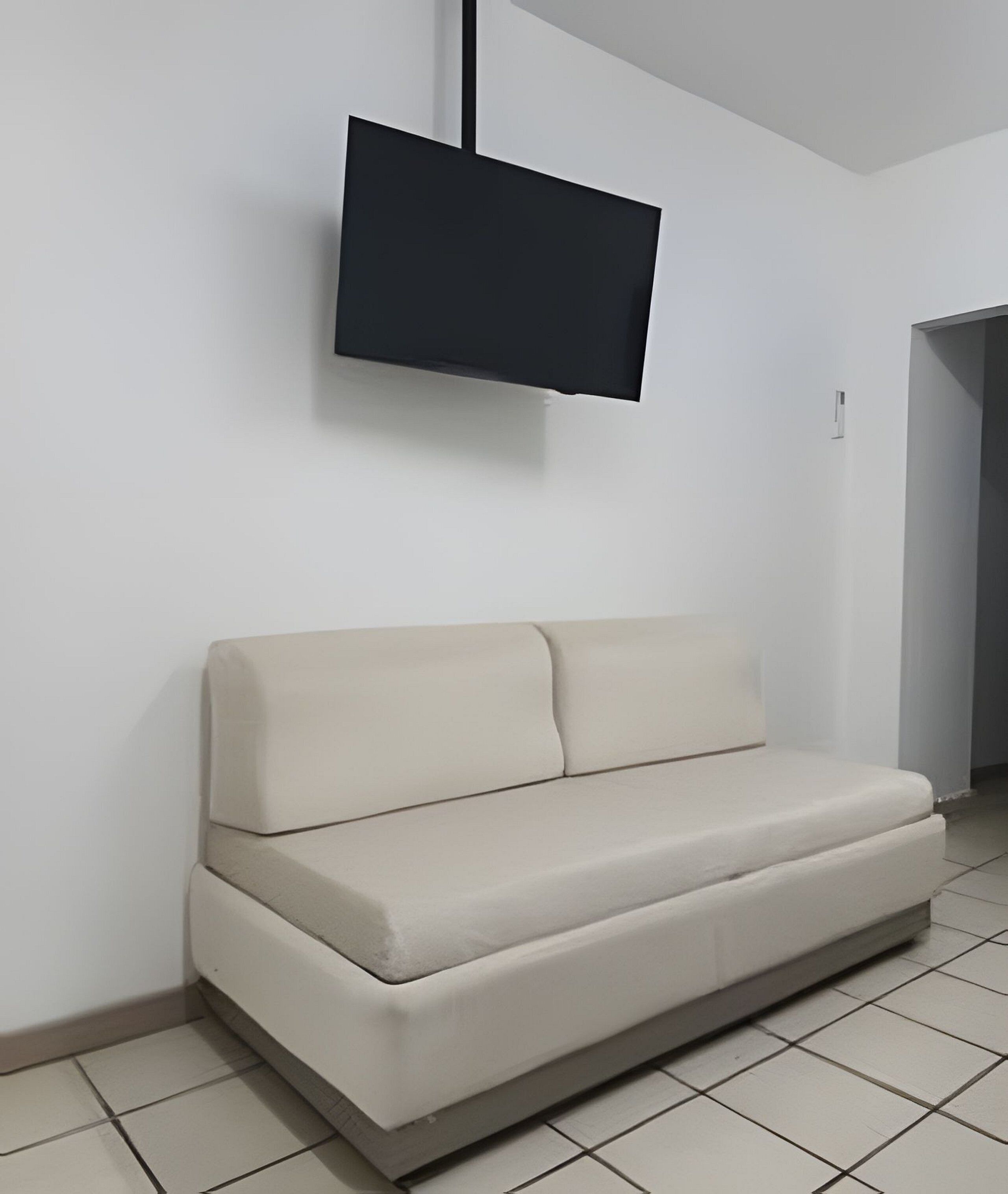 Una habitación con un sofá color beige, un televisor de pantalla plana montado en la pared y piso de baldosas.