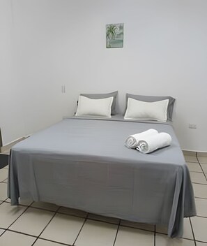 A bed with a grey bedspread and pillows, a small painting on the wall, and two rolled towels on the bed.