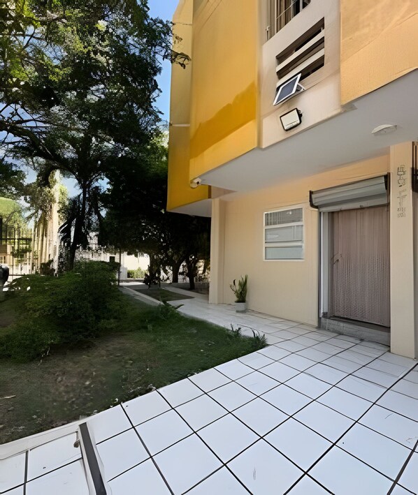 A building with a tiled entrance, a closed garage door, and a small garden area with trees and shrubs.