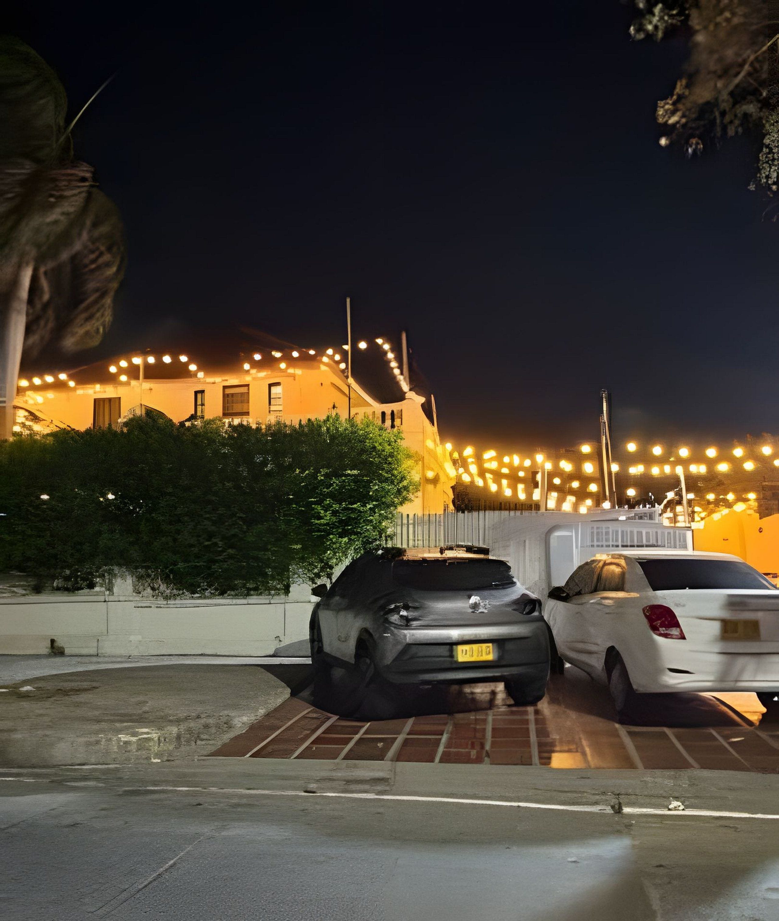 A parking area with two cars, a building with lights, and a fence.