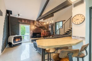 A modern living room with a fireplace, a dining area with a wooden table, and a staircase. - Notre écrin vosgien - Belle maison avec jacuzzi (Plainfaing)