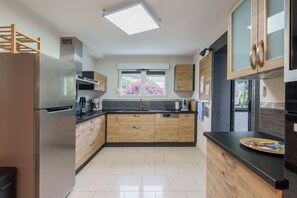 A modern kitchen with a stainless steel refrigerator, wooden cabinets, a black countertop, and a window with a view of trees. - Notre écrin vosgien - Belle maison avec jacuzzi (Plainfaing)