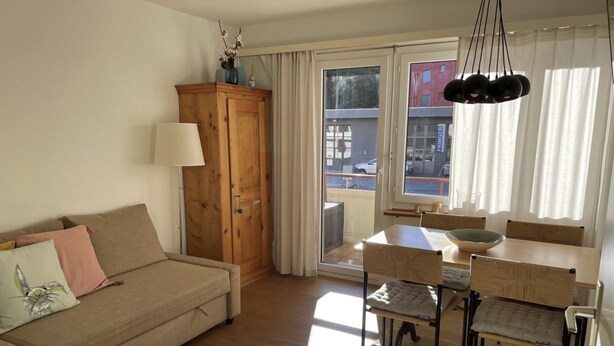 A furnished living room with a sofa, a wooden cabinet, a dining table with chairs, and a view through a glass door. - Paradies 017 Appartement/Fewo,   4-Bettwohnung by Interhome (Arosa)