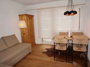 Een kamer met een houten vloer, een beige bank, een eettafel met stoelen, een houten kast en een raam met gordijnen.