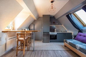 Une cuisine compacte avec un coin repas, un lit agrémenté d’un oreiller violet et une lucarne.