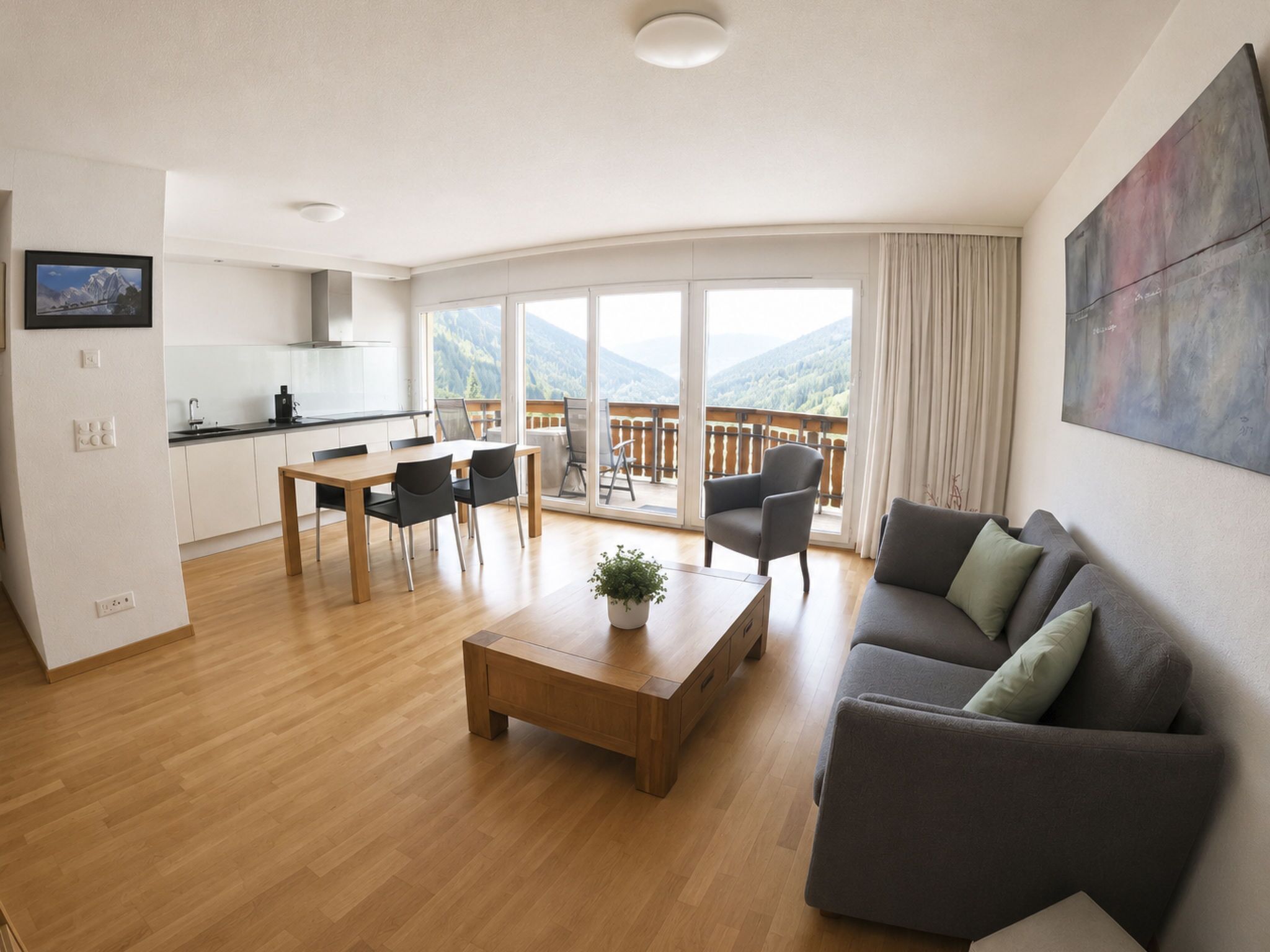A modern living room with a wooden coffee table, a grey sofa, and a balcony with a mountain view.