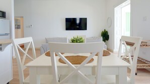 Un soggiorno moderno con televisione a schermo piatto, arredi bianchi e un'area pranzo con sedie bianche e un sottopiattino intrecciato.