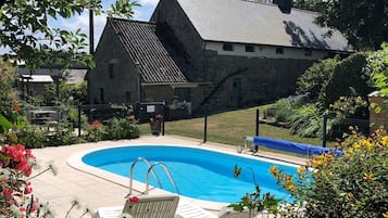A swimming pool surrounded by greenery, a stone building with a sloped roof, and a clear sky.