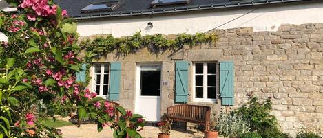 A stone house with a door, two windows with shutters, a bench, and a garden with flowers.