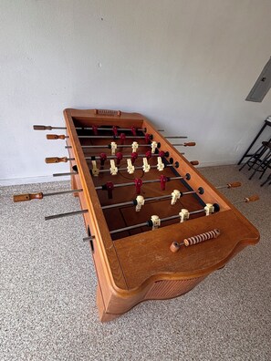 Ein hölzerner Kicker mit mehreren Stangen und Spielfiguren steht auf einem Teppichboden vor einer schlichten Wand.