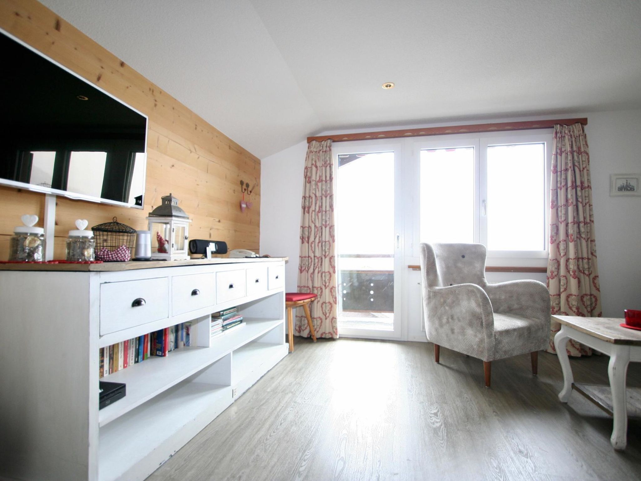 Een kamer met een houten wand, een witte kast met lades, een stoel, een tafel en een raam met gordijnen.