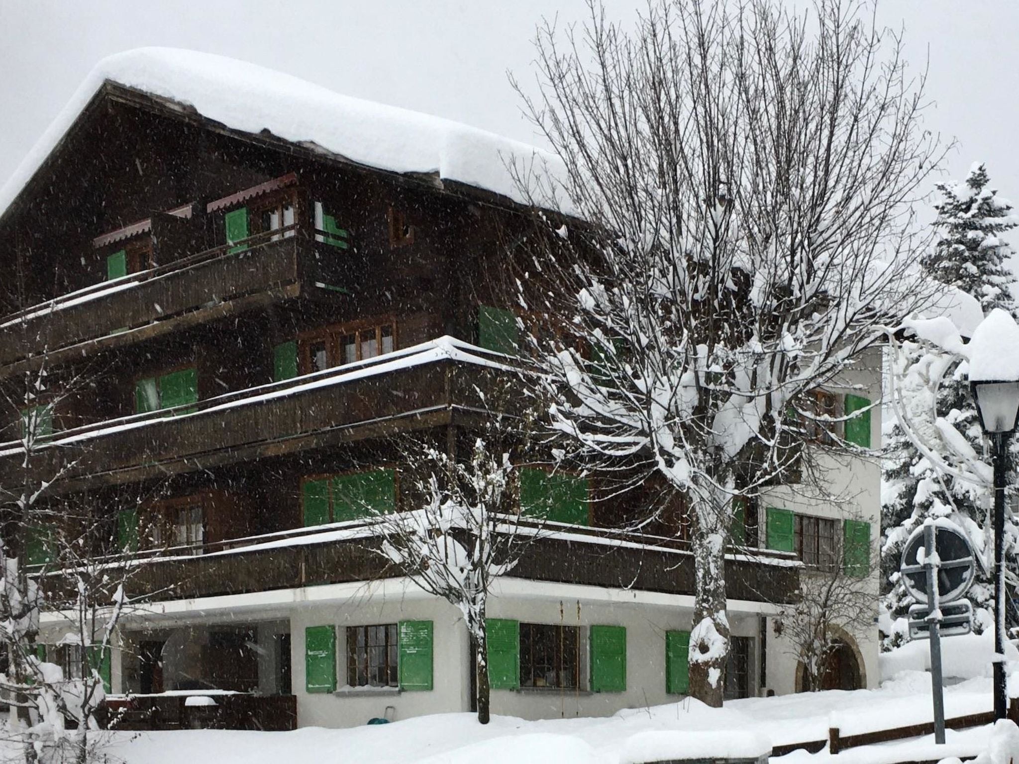 A multi-story building with a sloped roof covered in snow, surrounded by bare trees and a snow-covered street.