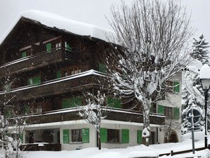 Ein mehrstöckiges Gebäude mit geneigtem, schneebedecktem Dach, umgeben von kahlen Bäumen und einer verschneiten Straße.