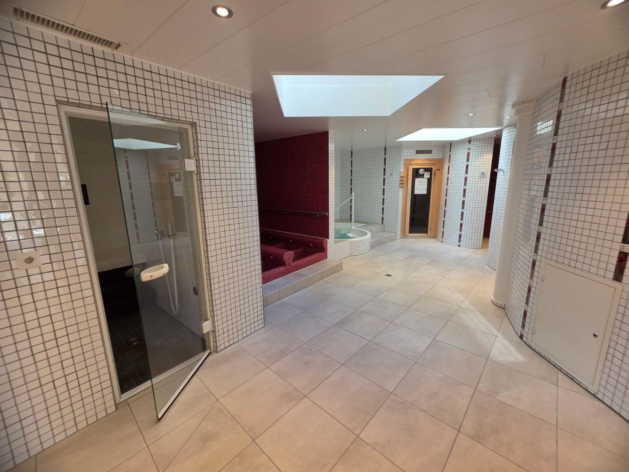 Un pasillo de hotel con paredes de azulejos, una puerta de cristal, una zona de estar roja y un tragaluz.