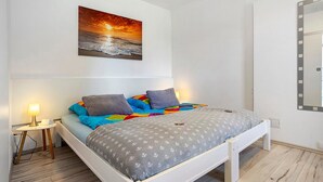 A bedroom with a bed, a bedside table with a lamp, a wall-mounted mirror, and a sunset painting. - Apartment 'Villa Wiese  Vineta' with Wi-Fi (Katzow)
