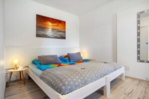 Ein Schlafzimmer mit einem Bett, einem Nachttisch mit Lampe, einem wandmontierten Spiegel und einem Sonnenuntergangsbild.