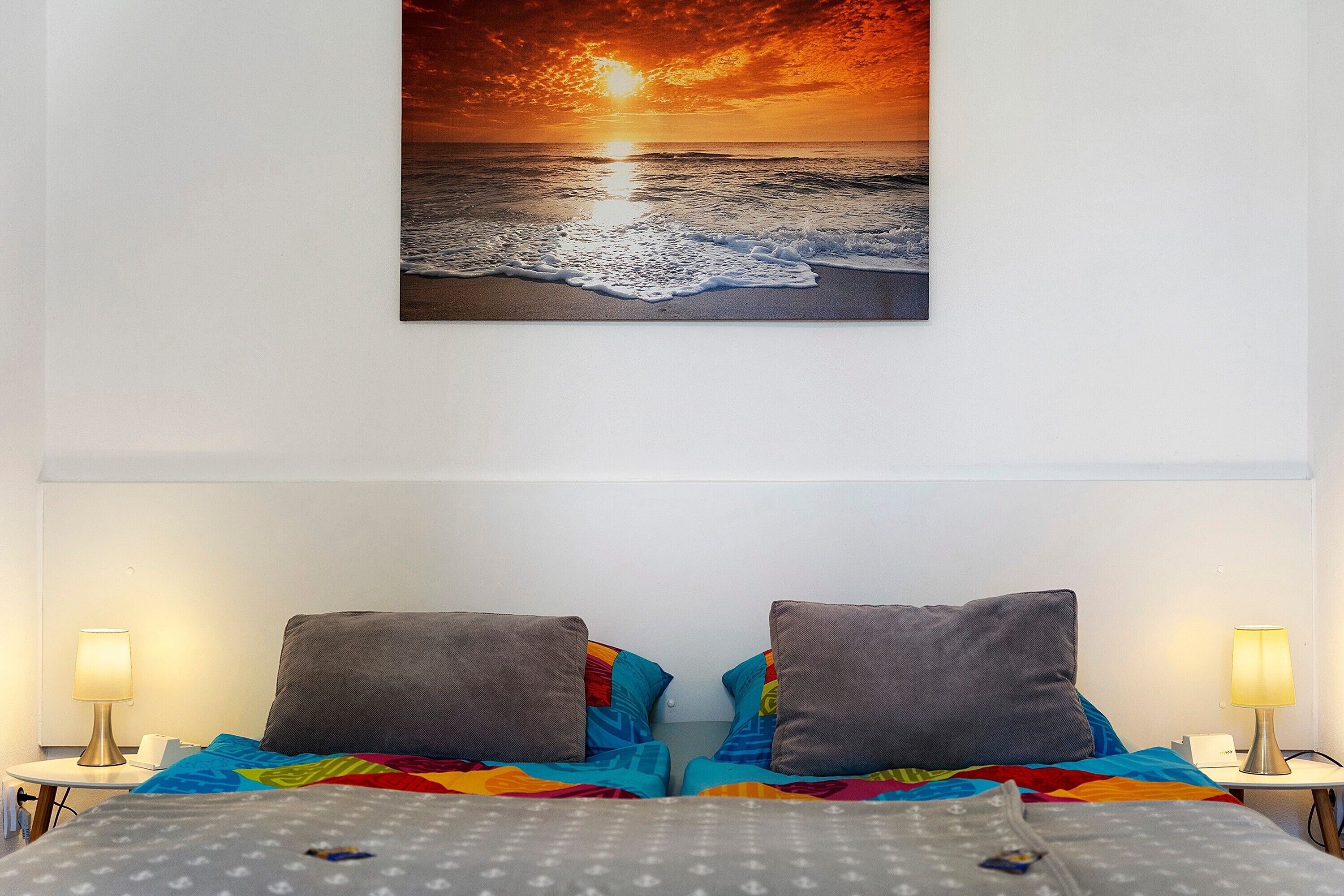 A hotel room with a bed, two pillows, a colorful blanket, a nightstand with a lamp, and a wall-mounted painting of a sunset over the ocean.