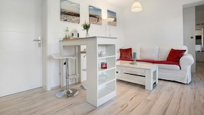 A modern living room with a white sofa, a coffee table, and a bookshelf. There are two paintings on the wall, a lamp, and a door to the left. - Apartment 'Villa Wiese  Vineta' with Wi-Fi (Katzow)
