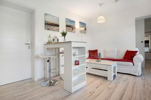 Ein modernes Wohnzimmer mit einem weißen Sofa, einem Couchtisch und einem Bücherregal. An der Wand hängen zwei Gemälde, eine Lampe und links eine Tür.