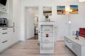 Et moderne køkken med hvide skabe, en indbygget ovn og en kaffemaskine. Der er et hvidt reolsystem med dekorative genstande og et rødt lysestage på et lille bord.