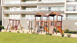 Un parque infantil con columpios y estructuras para escalar frente a un edificio moderno con balcones.