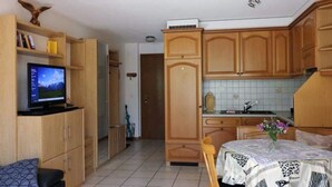 Un espacio habitable compacto con kitchenette, comedor con mesa y sillas, televisor en estante y armarios de madera.