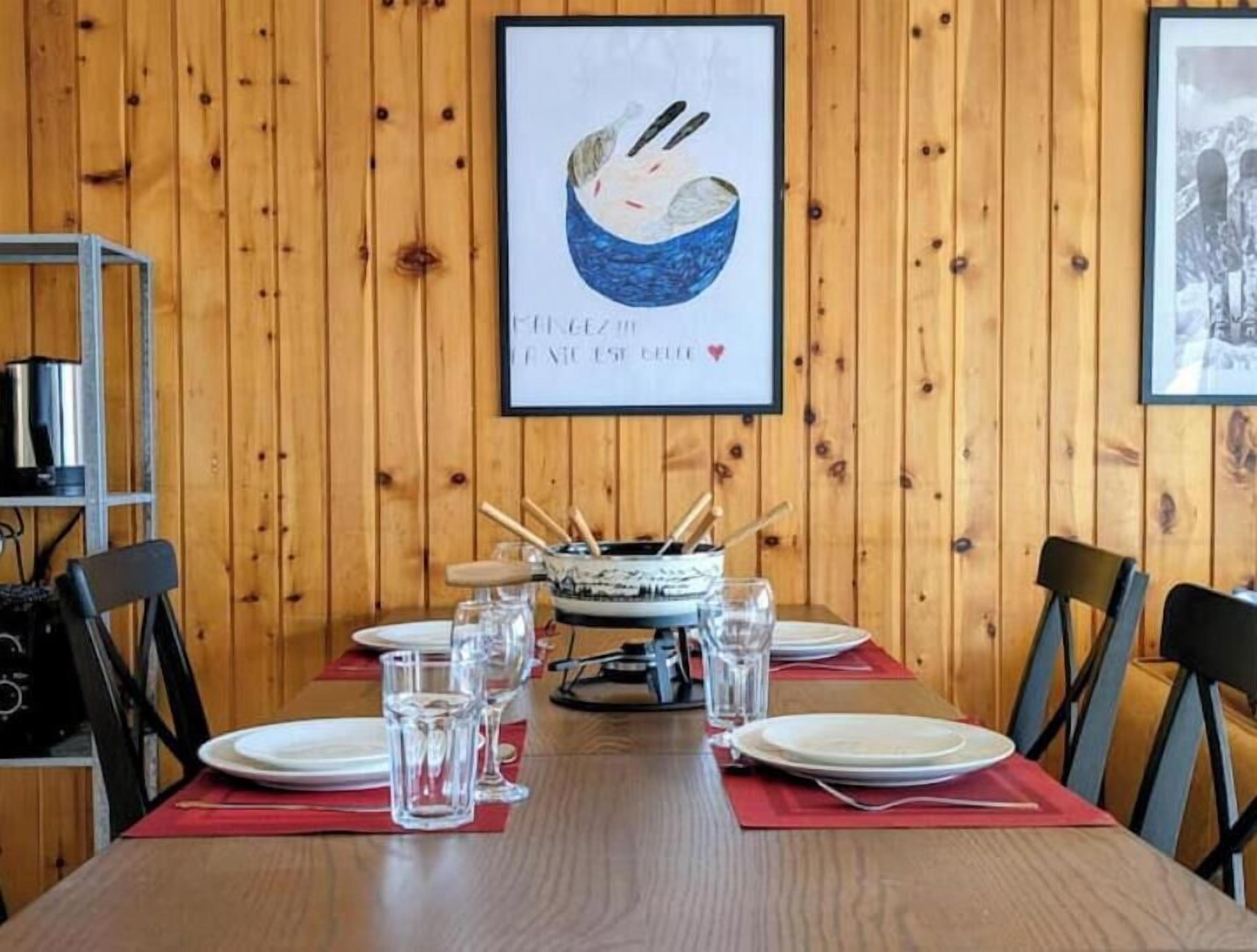 A dining table set with plates, glasses, and a fondue pot, with wooden paneling on the walls and a framed picture above.