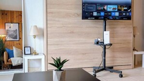 Moderni hotellihuone, jossa on seinätelineeseen asennettu litteänäyttöinen televisio, pieni pöytä, jossa on ruukussa oleva kasvi, valkoinen yöpöytä ja puinen seinäpaneeli.