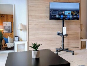 Ein modernes Hotelzimmer mit einem an einem Stand befestigten Flachbildfernseher, einem kleinen Tisch mit einer Topfpflanze, einem weißen Nachttisch und einer Holzvertäfelung.