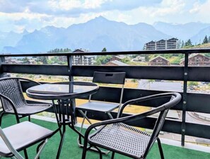Ein Balkon mit Tisch und Stühlen, der auf eine gebirgige Landschaft mit Gebäuden in der Ferne blickt.