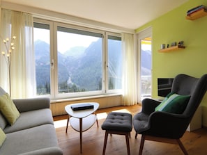Un salon comprenant un canapé, une petite table et un fauteuil, un téléviseur, et une vue sur les montagnes grâce à de grandes fenêtres.