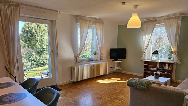 Ein Wohnzimmer mit Esstisch, Sofa, Fernseher, Bücherregal und großen Fenstern mit Vorhängen.