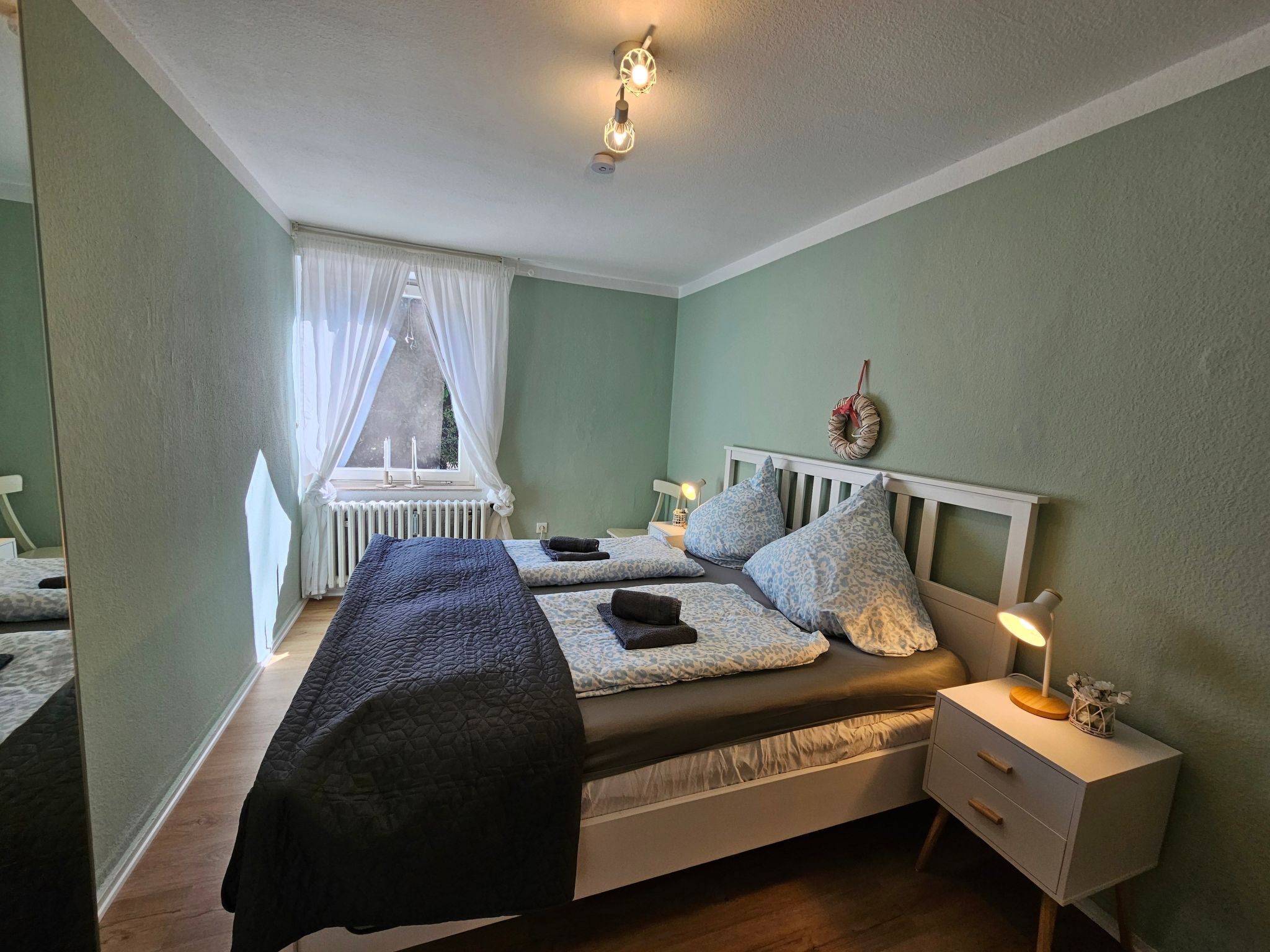 A bedroom with two beds, a bedside table with a lamp, a radiator, and a window with curtains.