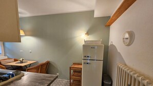 Eine kleine Küche mit einem Kühlschrank, einem Esstisch mit Stühlen, einem Heizkörper und einer Wanduhr.