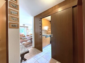 Un couloir avec une porte en bois donnant sur un espace cuisine, une table à manger avec des chaises, et un canapé dans le salon adjacent.