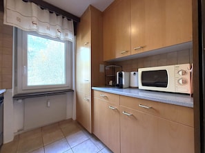 A kitchen with wooden cabinets, a window with a curtain, and a microwave on the countertop. - Lärchenwald 1808 # 4-bed apartment, Bad, WC 4-Bettwohnung, Bad, WC by Interhome (Leukerbad)