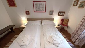 Una cama doble con ropa de cama blanca, dos mesitas de noche con lámparas, un banco y cuadros en la pared.